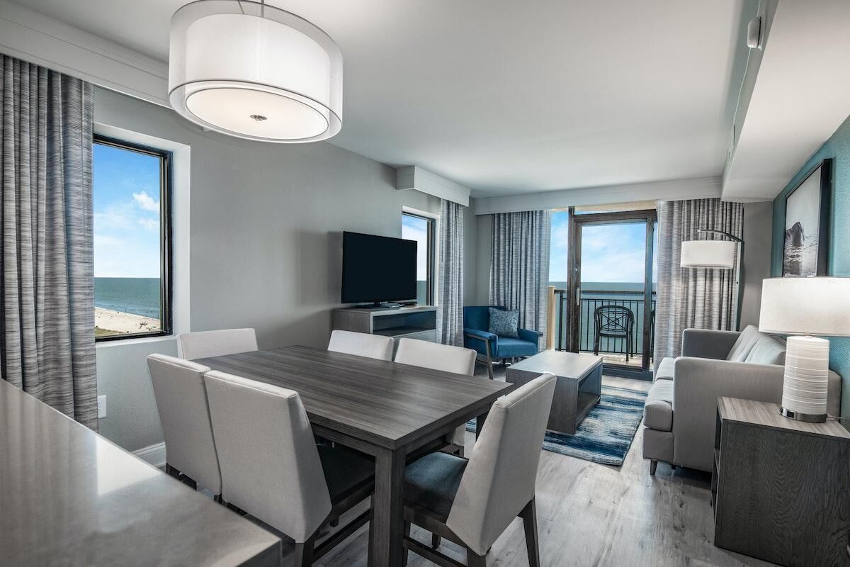 A modern hotel room with a dining area, a TV, a balcony with a view of the beach, and a seating area with a lamp.