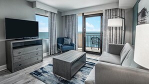 Una habitación de hotel moderna con televisión de pantalla plana, un sofá gris, un sillón azul y un balcón con vista al mar.
