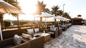 Un espace extérieur aménagé avec des canapés en osier et des parasols blancs, des palmiers et un sol carrelé.