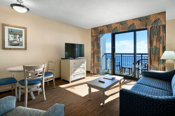 A hotel room with a balcony, a television, a dining area with chairs, a sofa, and a table.