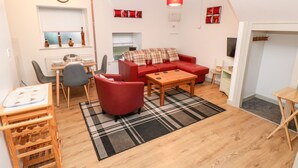 A compact living space with a red sofa, a wooden coffee table, a dining area with chairs, a kitchen area with a window, and a red pendant light.