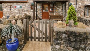 A stone cottage with a wooden gate, a bench, and a small tree in the garden.