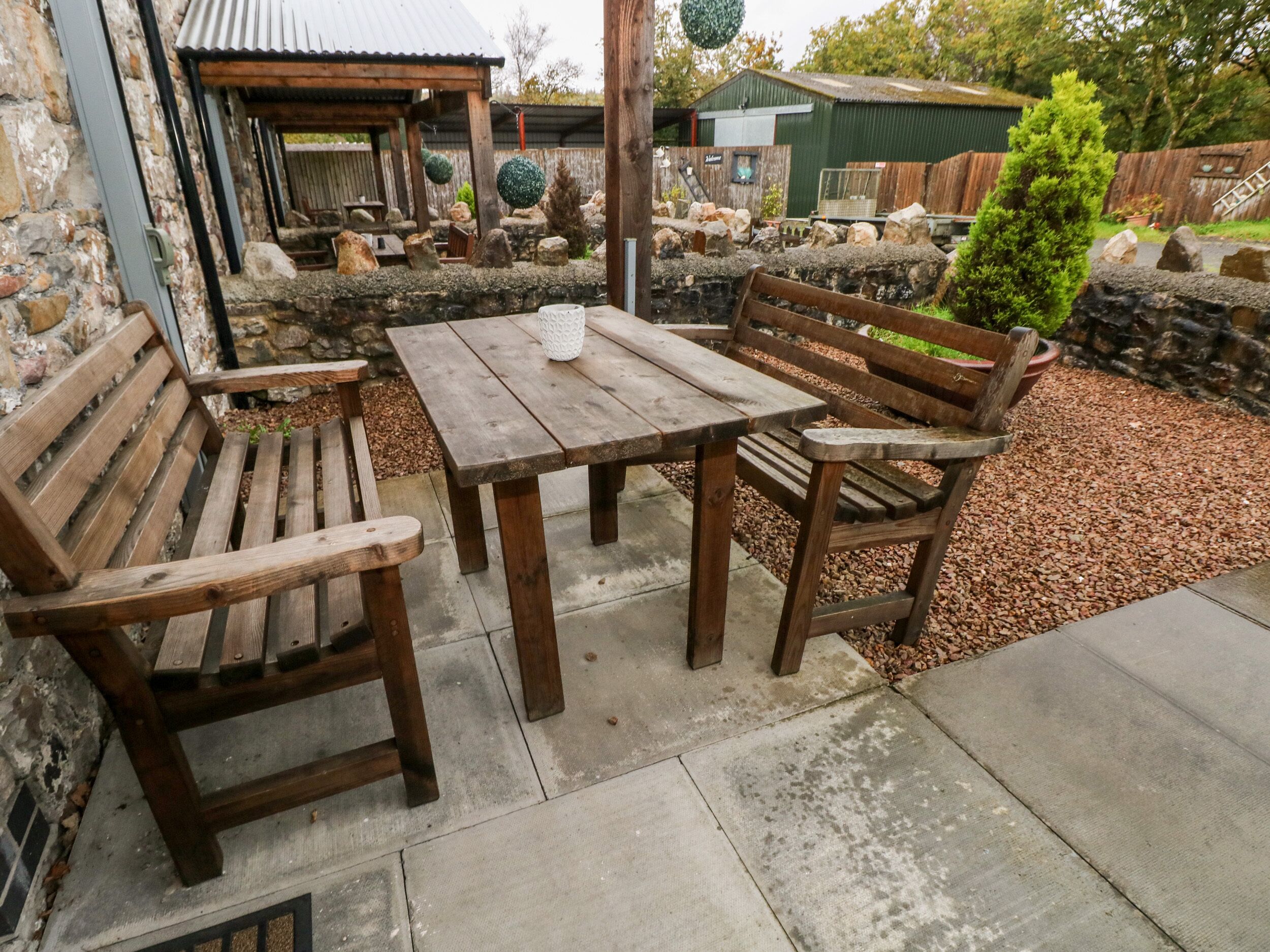 A wooden patio with a table and benches, surrounded by a stone wall and a small tree.