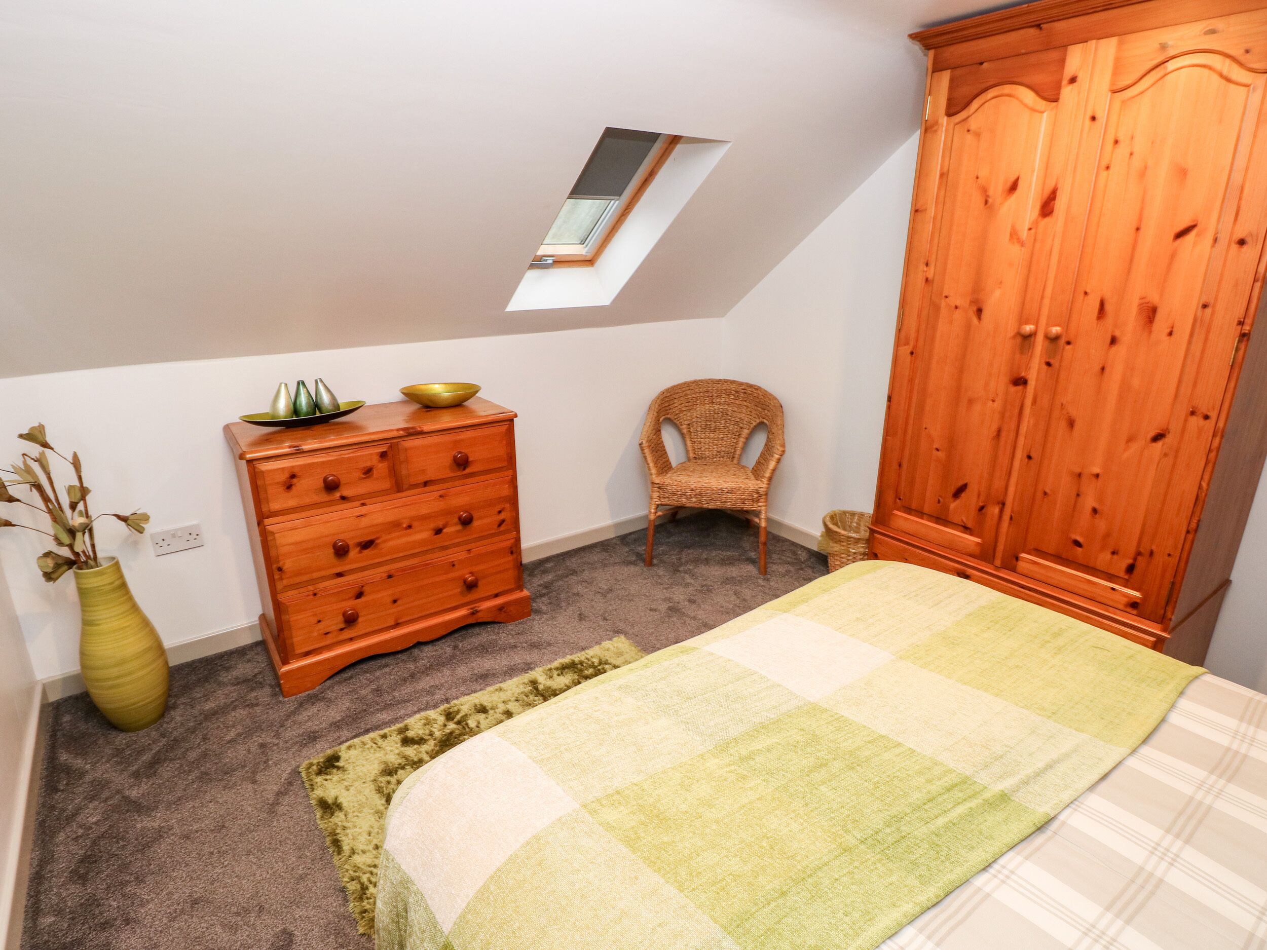 A bedroom with a bed, a wooden dresser, a wicker chair, a wardrobe, and a skylight.