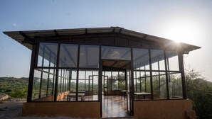 A glass-enclosed structure with a roof, featuring a dining area with tables and chairs, set against a hilly landscape. - Beyond Stay Jawai Legacy (Shivganj)