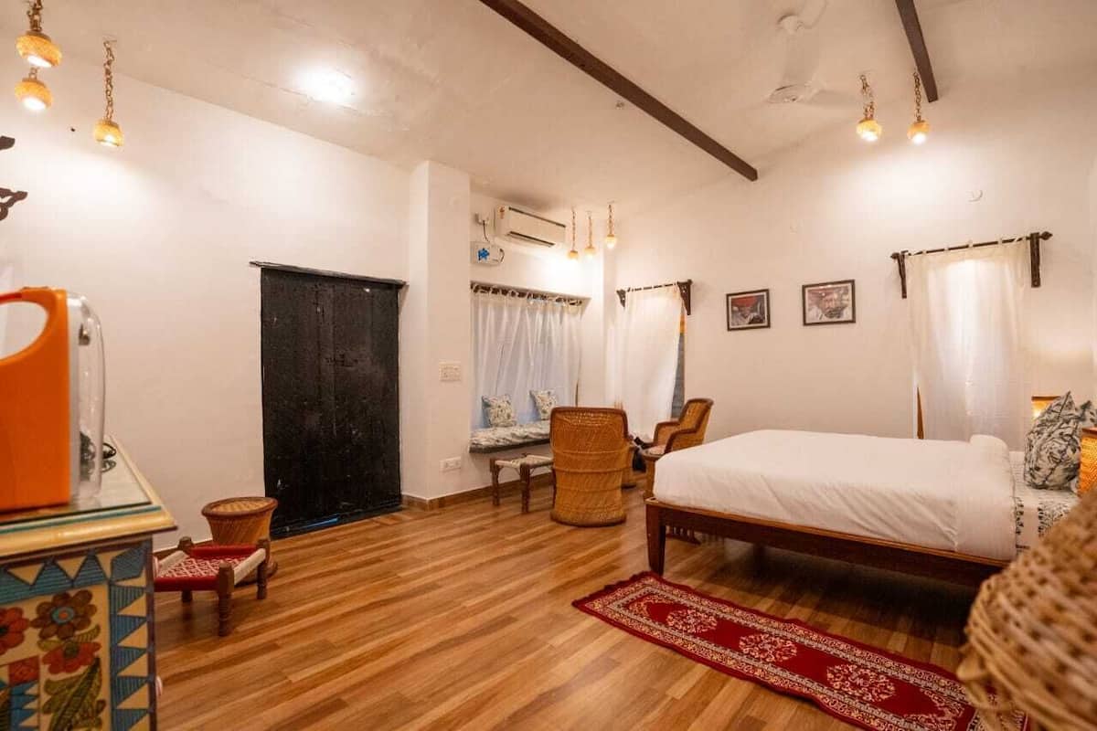 Un dormitorio con cama, piso de madera, alfombra roja, una silla, una mesa con tetera y una ventana con cortinas.