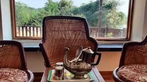 A room with a view of greenery through a window, a wicker chair, a table with a teapot and cups, and another wicker chair. - Beyond Stay Jawai Legacy (Shivganj)