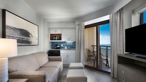 Ein modernes Hotelzimmer mit einer Couch, einem Fernseher, einem Esstisch mit Stühlen, einer Küchenzeile und einem Balkon mit Blick.