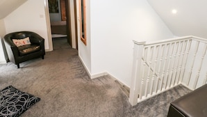 Un escalier avec des rampes blanches mène au deuxième étage. Un fauteuil en cuir noir, agrémenté d’un coussin, est placé près du bas de l’escalier.