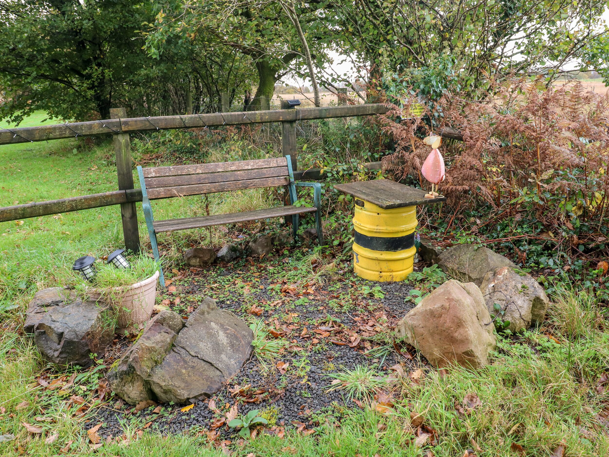 A wooden bench, a yellow barrel, and a pink flamingo statue in a garden with rocks and foliage.