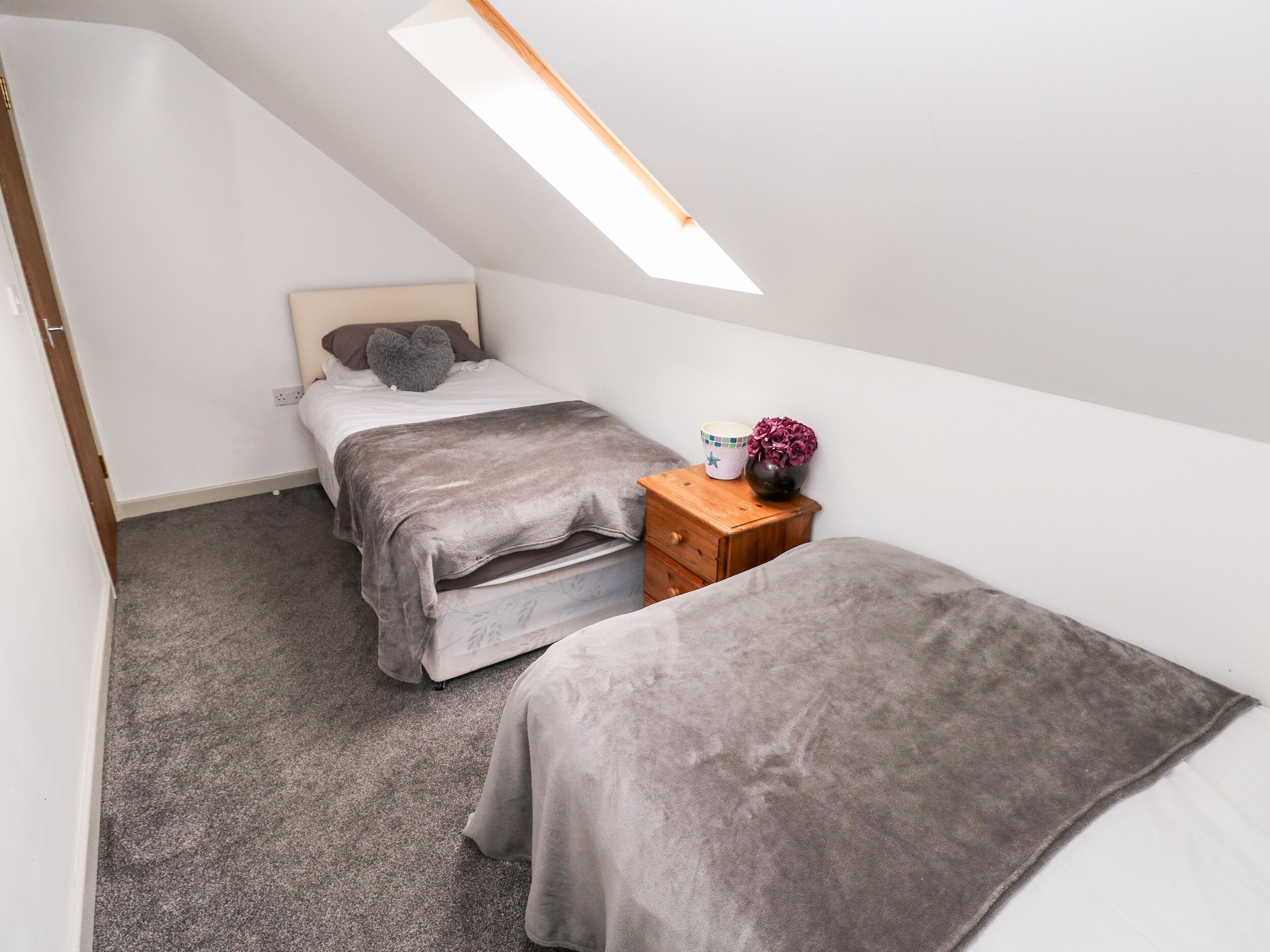 A small, modern bedroom with a sloped ceiling and a skylight. There are two beds with grey bedding, a wooden nightstand with a vase of flowers, and a white wall.