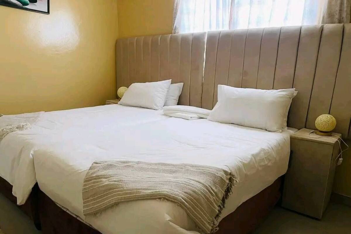 Una cama bien hecha con sábanas blancas y una manta color beige, un cabecero de madera y una lámpara de noche con base esférica.