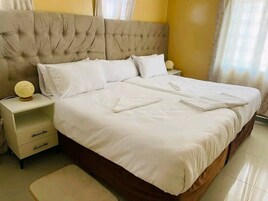 Una cama bien tendida con sábanas y almohadones blancos, un cabecero capitonado y una lámpara de noche sobre una mesita.