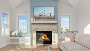 Una sala de estar con chimenea, un televisor grande montado encima y vistas al exterior a través de amplias ventanas.