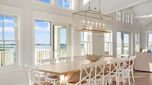 Un comedor amplio con una gran mesa de madera y sillas blancas, ventanas grandes que ofrecen vistas al mar y una lámpara moderna.