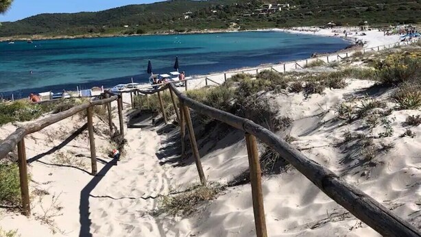 A sandy beach with a wooden staircase leading down to the water, a clear blue sky, and a distant view of a hilly landscape with buildings. - 3 bedroom villa in sardinia (Sardegna)