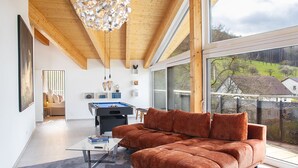 A modern living room with a brown sofa, a pool table, and a large window offering a view of the outdoors.