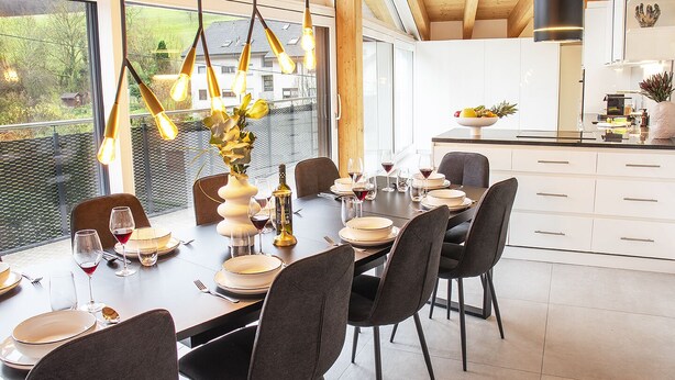 A dining area with a long table set for six, surrounded by chairs. There is a vase with flowers on the table, and a bottle of wine is visible. The room has large windows, a kitchen in the background, and a ceiling with wooden beams.