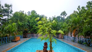 Piscina con una estatua en el centro, rodeada de exuberante vegetación y una cerca azul.