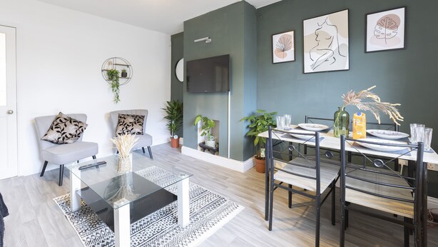 Een moderne woonkamer met een glazen salontafel, grijze banken en een eethoek met een houten tafel en metalen stoelen.