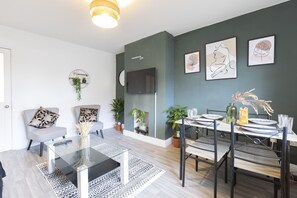 A modern living room with a glass coffee table, grey sofas, and a dining area with a wooden table and metal chairs. - Modern Cosy 3BR Home Sleeps 7 Free WiFi & Parking (Osmondthorpe)