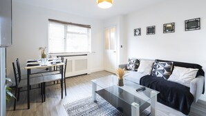 Een moderne woonkamer met een glazen salontafel, een bank met opvallende kussens, een eettafel met stoelen en een groot raam met zonwering.
