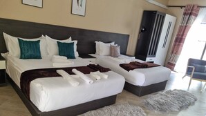 A hotel room with two beds, a large dark headboard, a grey furry rug, and a grey furry ottoman. - Eden Wood Guest House (Shakawe)