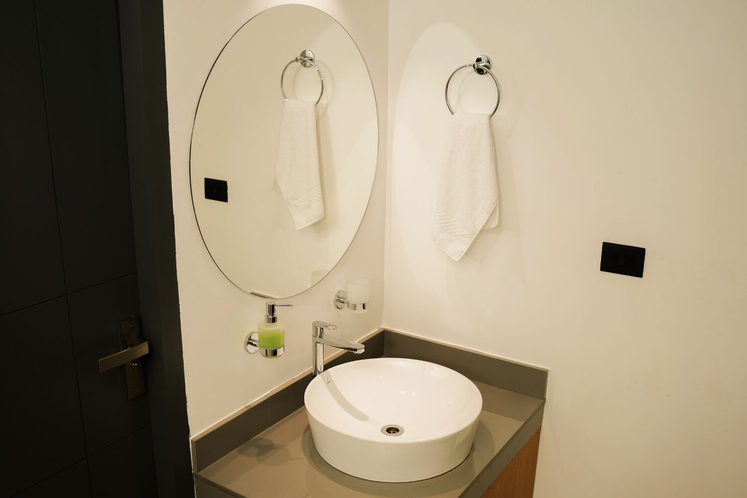 A modern bathroom with a round sink, two towel rings, a mirror, and a soap dispenser.