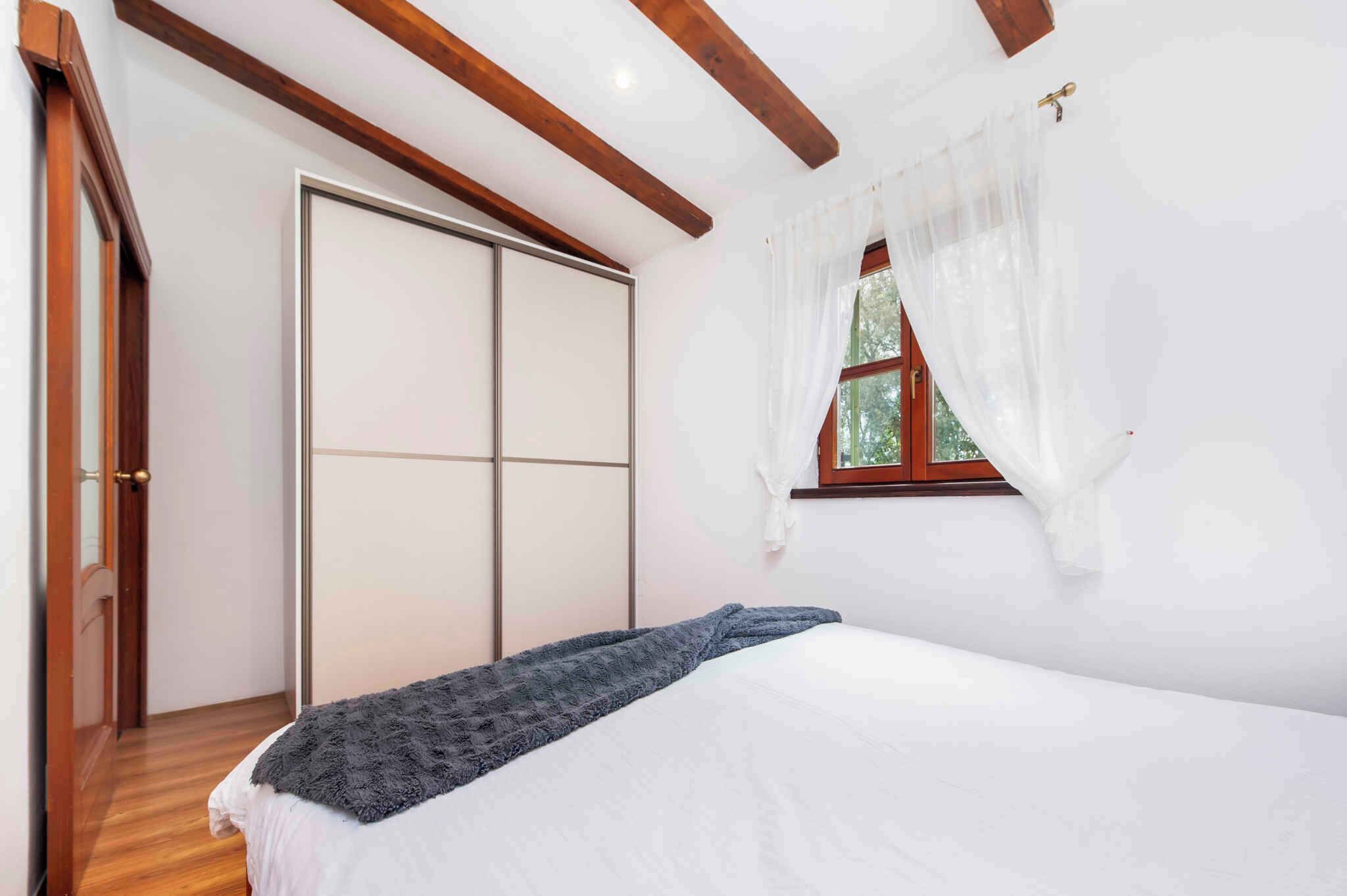 A bedroom with a bed, a wardrobe, a window with curtains, and wooden beams on the ceiling.