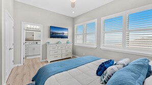 Un dormitorio con cama, tocador con espejo, televisor y ventana con persianas.