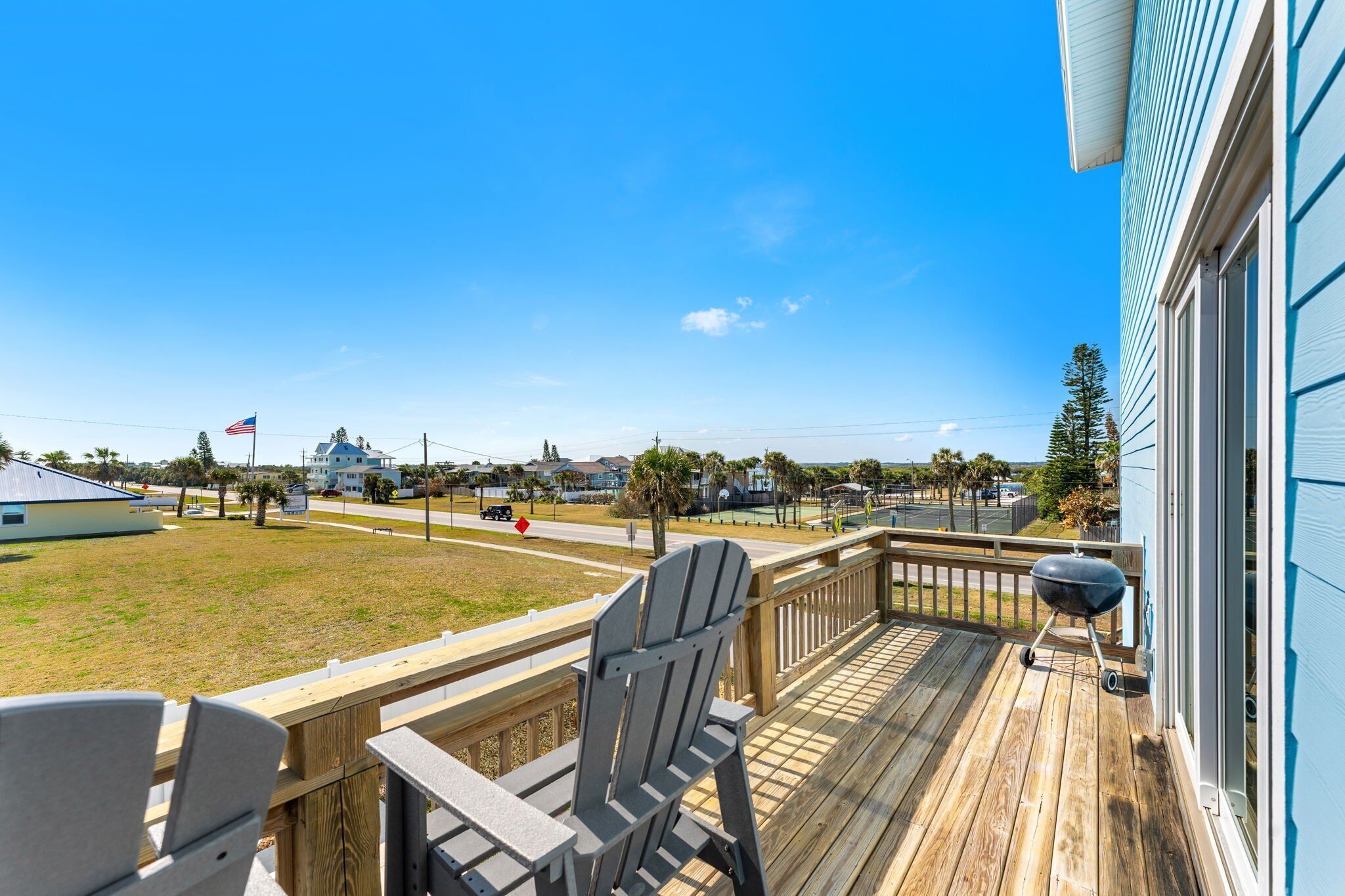 Una terraza de madera con sillas Adirondack, una parrilla y vista a una zona residencial con casas y una bandera.