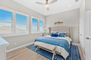 Une chambre à coucher comprenant un lit, un ventilateur de plafond, une table de chevet et une fenêtre avec des stores.