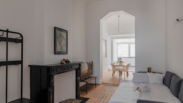 Een moderne hotelkamer met een houten vloer, een bed met een grijze hoofdeinde, een zwarte open haard, een zithoek met een tafel en stoelen, en een groot raam dat voor natuurlijk licht zorgt.