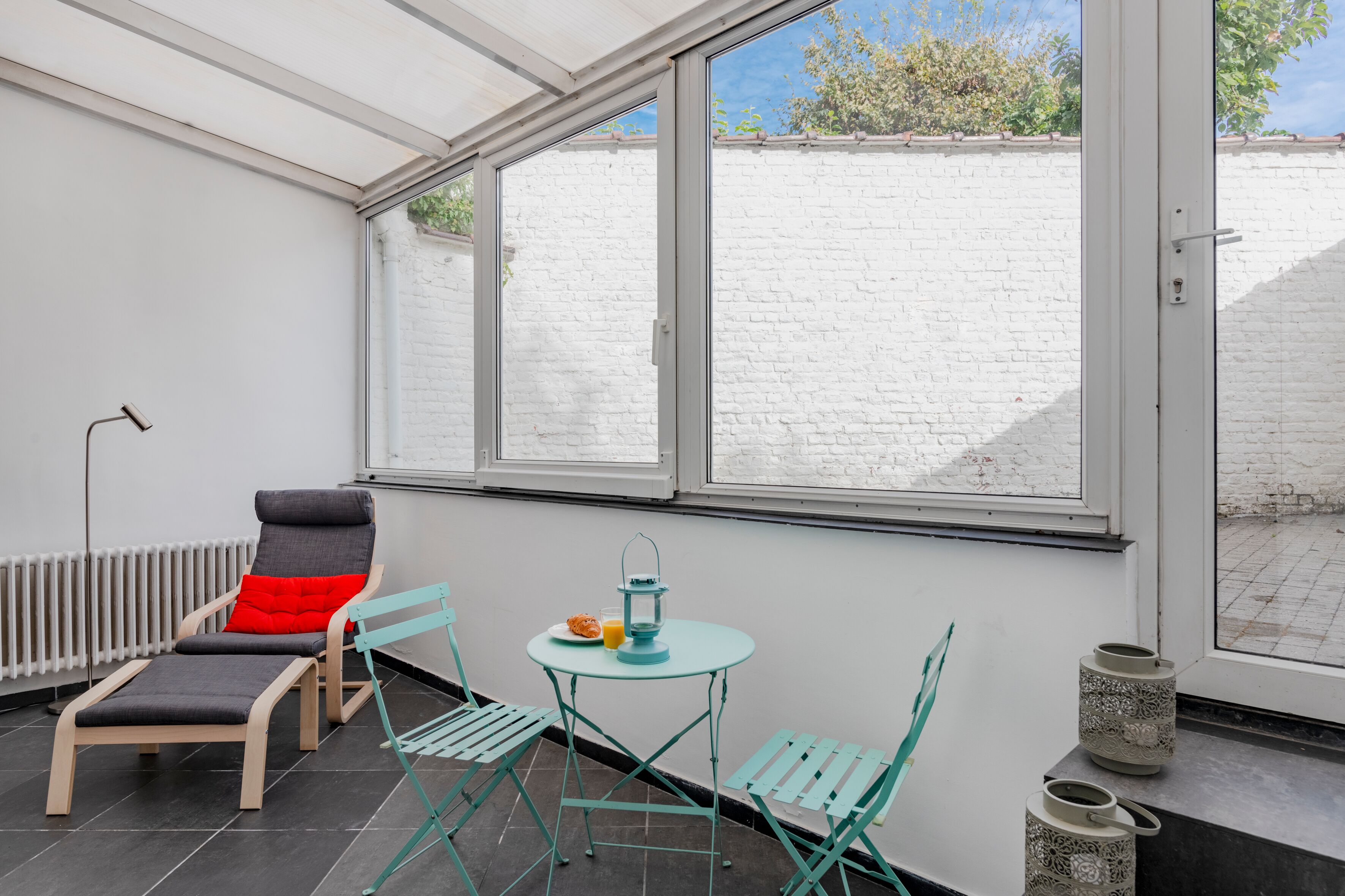 Une véranda avec un mur en briques blanches, un radiateur, une table avec des chaises et une fenêtre donnant sur un mur en briques et de la verdure.