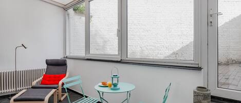 Une véranda avec un mur en briques blanches, un radiateur, une table avec des chaises et une fenêtre donnant sur un mur en briques et de la verdure.