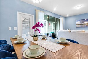 A dining area with a wooden table set for four, a vase with purple flowers, and a sofa in the background.
