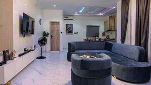 A modern living room with a grey sectional sofa, a small ottoman, a flat-screen TV mounted on the wall, a standing fan, and a door leading to the kitchen. - RHEMA APARTMENTS (Lagos)