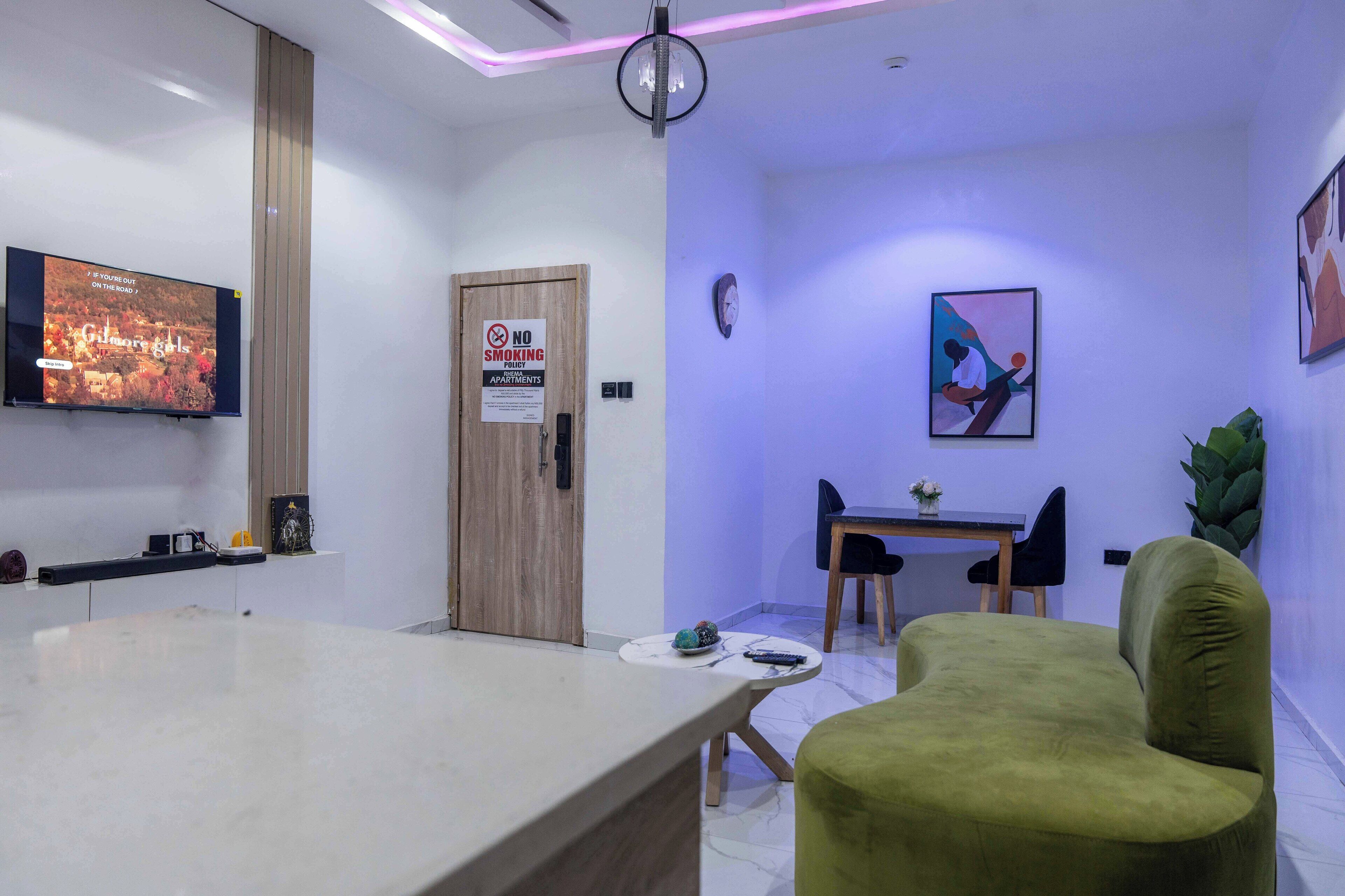 A modern living room with a flat-screen TV, a wooden door, a dining area with a table and chairs, and a green armchair.