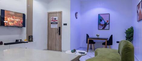 Una sala de estar moderna con televisor de pantalla plana, una puerta de madera, un comedor con mesa y sillas, y un sillón verde.