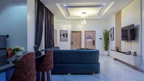A modern living room with a dark blue sofa, a dining area with two chairs, a TV mounted on the wall, and a ceiling with recessed lighting. - RHEMA APARTMENTS (Lagos)