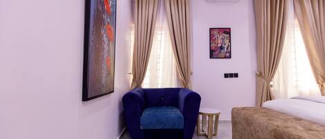 Una habitación con un sillón azul, una mesita redonda pequeña, una cama, cortinas color crema y dos aires acondicionados instalados en la pared.