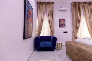 A room with a blue armchair, a small round table, a bed, beige curtains, and two wall-mounted air conditioners. - RHEMA APARTMENTS (Lagos)