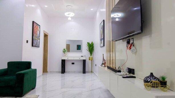 A modern living room with a green armchair, a white console table, a flat-screen TV mounted on the wall, and a small plant on the console table. - RHEMA APARTMENTS (Lagos)