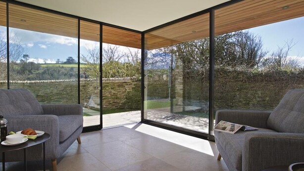 A modern room with large glass doors leading to a garden. There are two grey sofas and a small table with a plate and cup.