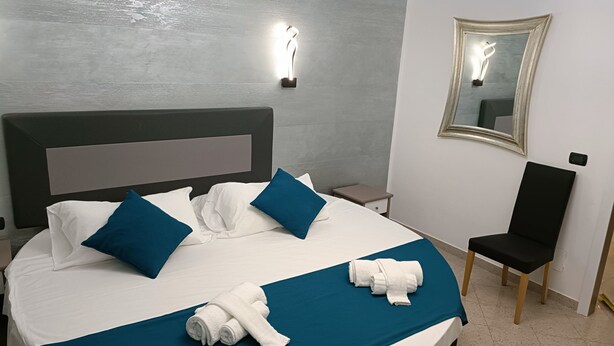 A neatly made bed with white and blue bedding, a teal bedspread, and rolled white towels. There is a small bedside table with a lamp, a black chair, and a mirror on the wall. - House Quattro Mori (Rome)