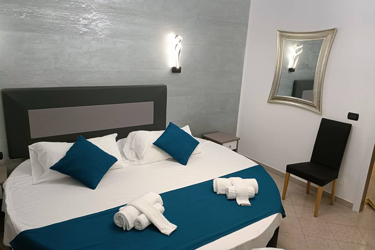 A neatly made bed with white and blue bedding, a teal bedspread, and rolled white towels. There is a small bedside table with a lamp, a black chair, and a mirror on the wall.