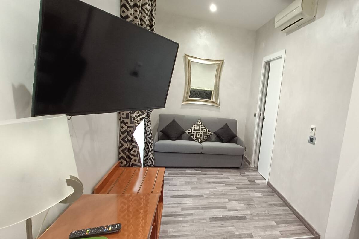 A modern living room with a flat-screen TV mounted on the wall, a grey sofa with patterned cushions, a wooden coffee table, and a patterned rug.