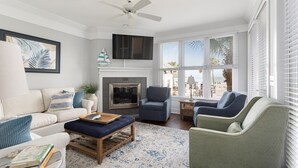 Un salon doté d’une cheminée, d’un ventilateur de plafond et offrant une vue sur la plage grâce à de grandes fenêtres.