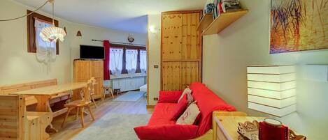 A cozy living area with a wooden sofa, a dining table with chairs, a TV, and a bookshelf.