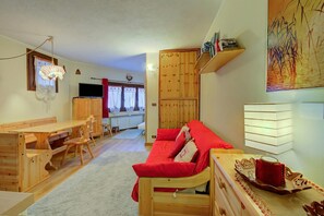 Un salon chaleureux avec un canapé en bois, une table à manger avec des chaises, un téléviseur et, au fond, une cuisine.